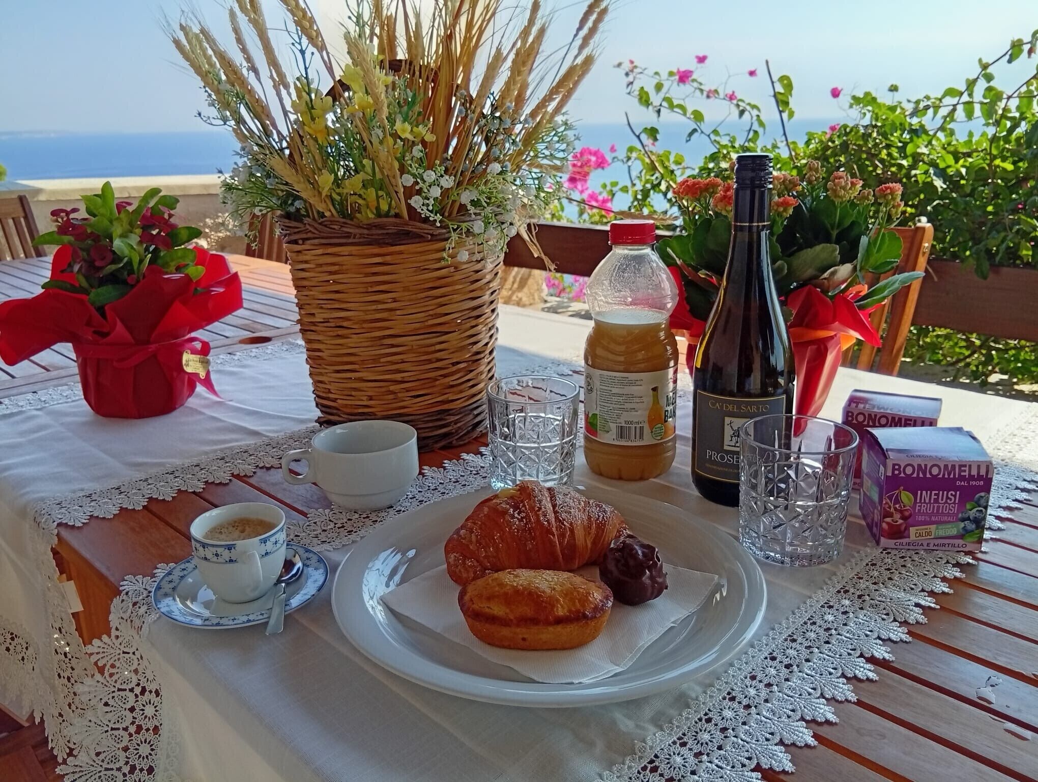 Une table dressée avec une bouteille de vin, un verre d’eau, une boîte de jus de fruits Bonomelli, un croissant, une pâtisserie et une tasse de café.