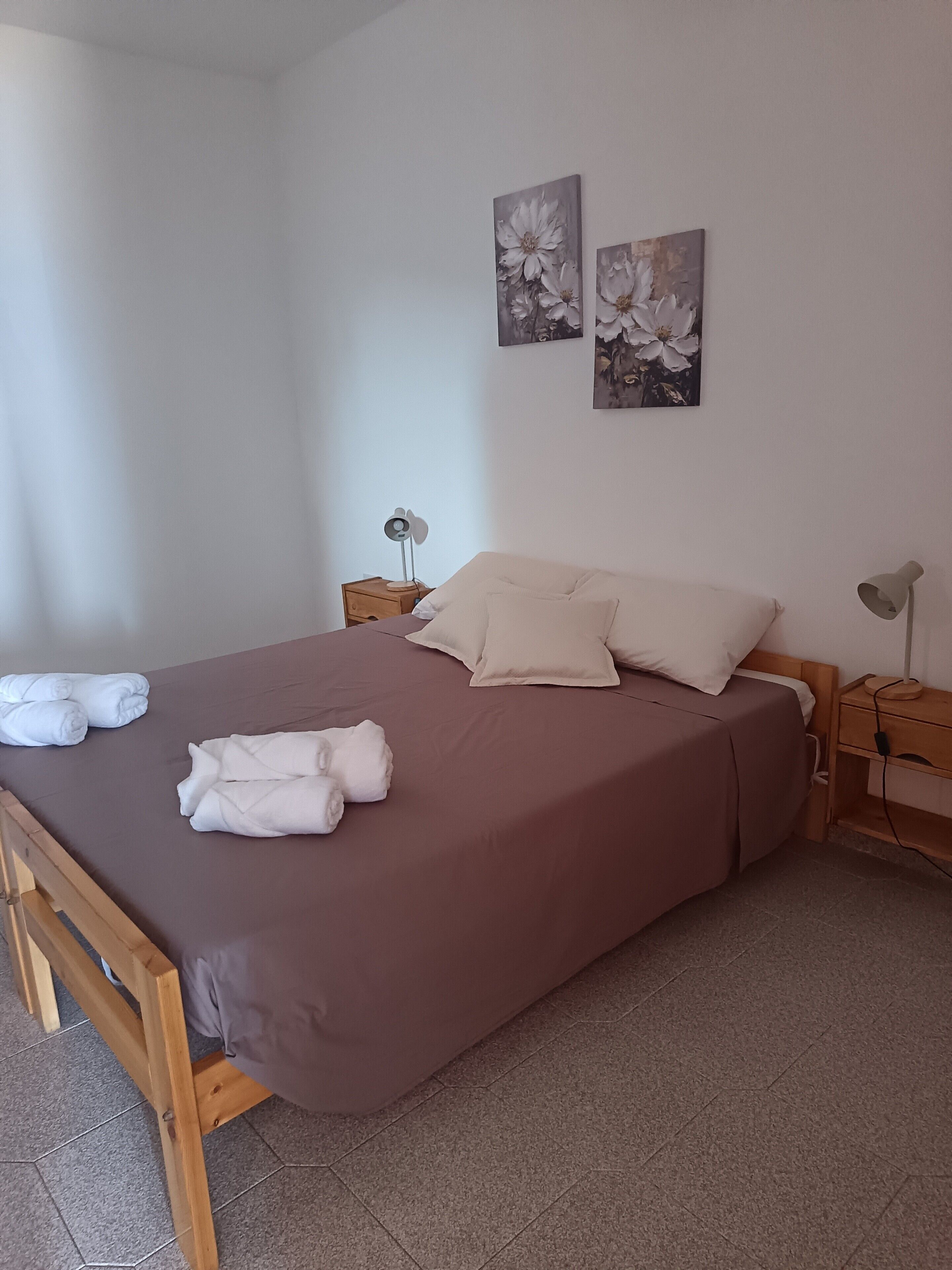 Un lit bien fait, agrémenté d’oreillers et de serviettes, une table de chevet avec une lampe, et deux tableaux représentant des fleurs, accrochés au mur.