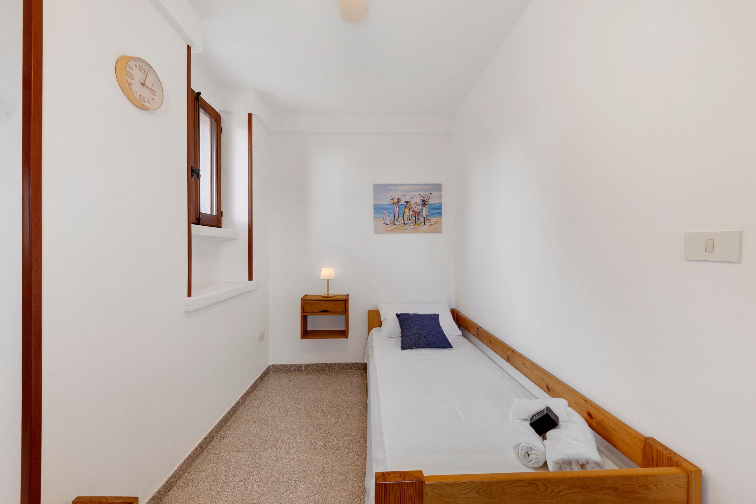 Ein kleiner, sauberer Raum mit einem Bett, einem Nachttisch, einer Wanduhr und einem Gemälde.