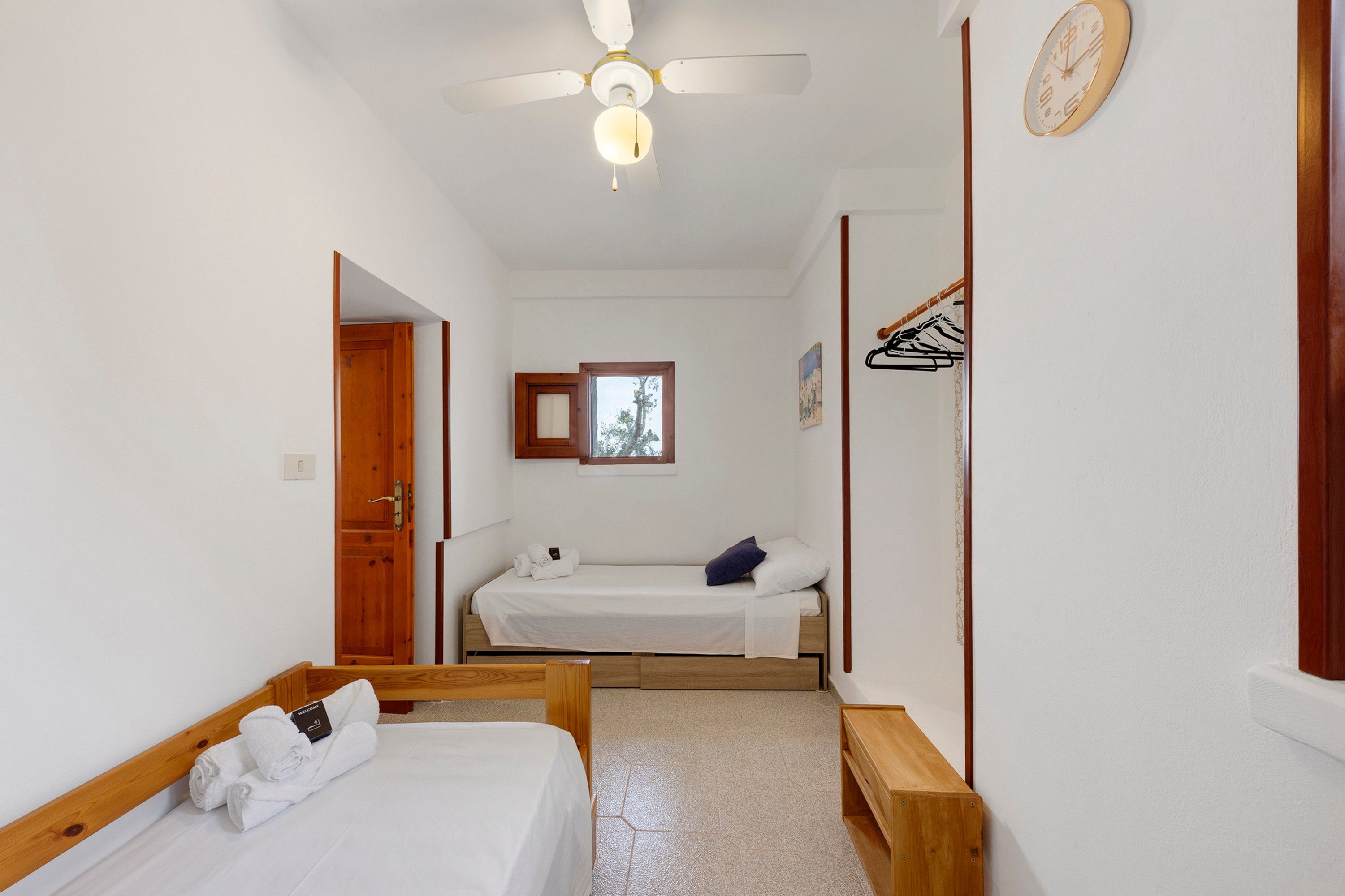 Habitació d'hotel amb un llit, una taula de nit, un ventilador de sostre, un rellotge i una finestra.