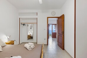 A bedroom with a bed, a bedside table, a lamp, a mirror, and a view of the sea through an open door.