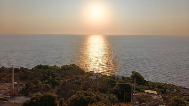 A coastal landscape with a sunset over the sea, a small settlement with houses, and a hill with vegetation.