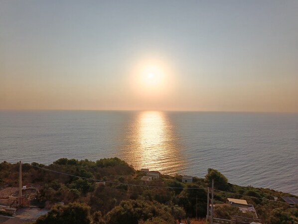 A coastal landscape with a sunset over the sea, a small settlement with houses, and a hill with vegetation.