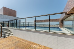 Una piscina en la azotea con barandillas de vidrio, terraza de madera y vista a la ciudad.