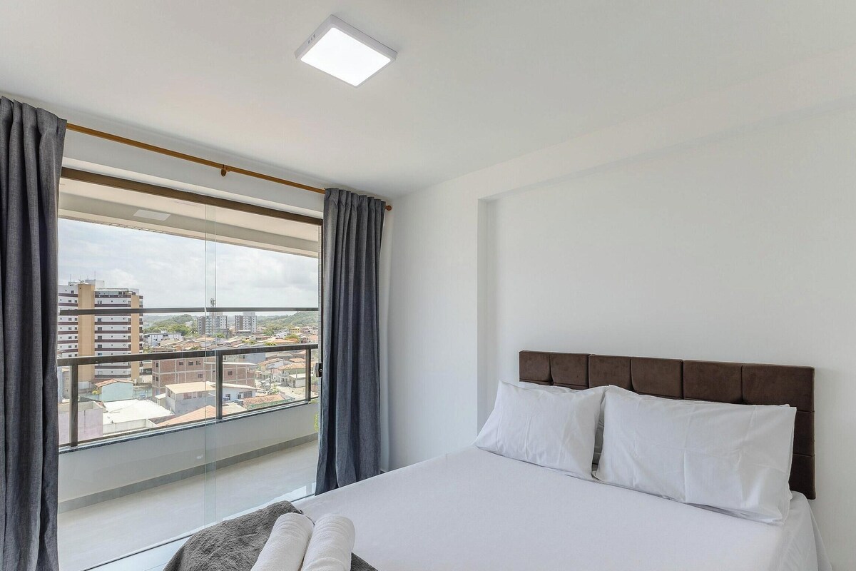 Quarto de hotel com uma grande janela, cama com roupa de cama branca e vista para a paisagem urbana.