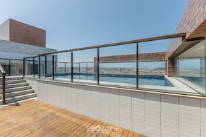 Piscina no terraço com guarda-corpos de vidro, deck de madeira e vista para a cidade.