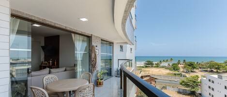 A balcony with a table and chairs, a view of the sea, and a glimpse of the surrounding buildings.