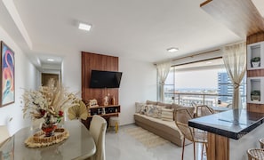 Una sala de estar moderna con sofá, mesa de comedor y sillas. Un televisor de pantalla plana está montado en la pared, y hay un ventanal con vista a la ciudad.