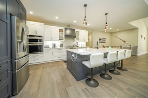 A modern kitchen with stainless steel appliances, a central island with bar stools, and a dining area with a view of the outdoors. - Executive Cottage on Lakeview HotTub Firepit Sauna Nearby Walking Nature Trails (Lambton Shores)