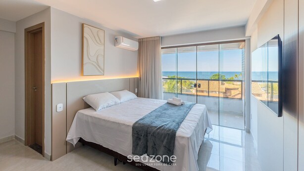 A modern bedroom with a bed, a ceiling light, a window with a view of the sea, and a mounted TV. - Sea View Apartment 300m From the Beach Mmd0403 (Ilhéus)