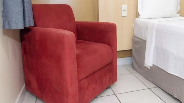 Un fauteuil rouge est placé à côté d’un lit recouvert de linge de lit blanc, dans une pièce dont un mur est en bois et le sol carrelé.