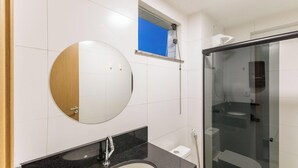 A modern bathroom with a round mirror, a glass shower enclosure, and a small window above the mirror. - Apt With Balcony and sea View in Ilheus - Mmd0503 (Ilhéus)