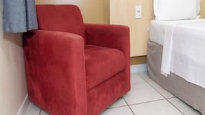 Un fauteuil rouge est placé à côté d’un lit recouvert de linge de lit blanc, dans une pièce dont un mur est en bois et le sol carrelé.