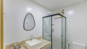 A modern bathroom with a glass-enclosed shower, a white sink with a marble countertop, and a large oval mirror. - Stay Near sea w/ Pool & Balcony Mmd0604 (Ilhéus)