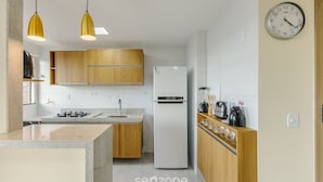 A modern kitchen with wooden cabinets, a white refrigerator, and a clock on the wall. - Stay Near sea w/ Pool & Balcony Mmd0604 (Ilhéus)