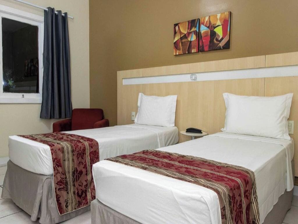 Ein Hotelzimmer mit zwei Einzelbetten, einem roten Sessel, einem kleinen Tisch und einem Fenster mit Vorhängen.