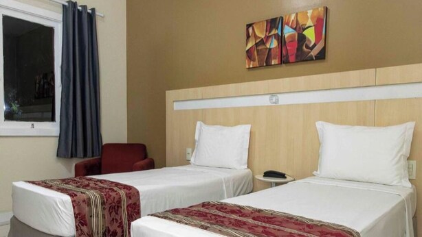 Ein Hotelzimmer mit zwei Einzelbetten, einem roten Sessel, einem kleinen Tisch und einem Fenster mit Vorhängen.