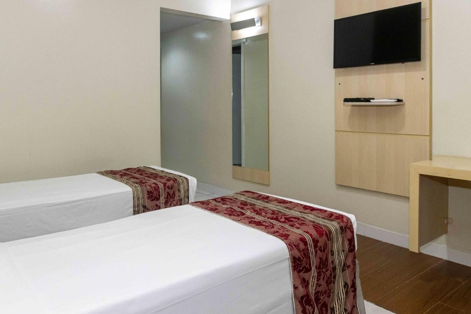 Ein Hotelzimmer mit zwei Einzelbetten, einem Spiegel, einem an der Wand befestigten Fernseher und einem kleinen Holztisch.