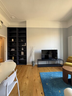Un salon moderne avec un plancher en bois, un meuble de rangement noir pour appareils électroniques, un vase blanc orné de branches, un téléviseur à écran plat posé sur un support noir et un canapé jaune.