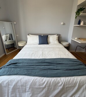 A neatly made bed with a white and blue duvet, two bedside tables, a mirror, and a small plant on a shelf. - La Dolce Vita - Vintage Two-Room Apartment in Dateo (Milano)