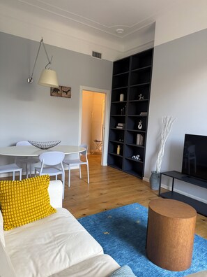Ein modernes Wohnzimmer mit einer Couch, einem Couchtisch und einer TV-Konsole. Es gibt einen Essbereich mit Tisch und Stühlen sowie ein Bücherregal voller Dekoration.