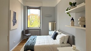 Ein modernes Schlafzimmer mit Bett, Nachttisch und Blick auf die Stadt durch ein Fenster.