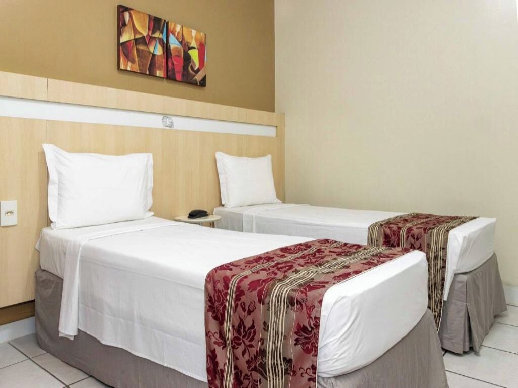 Ein Hotelzimmer mit zwei Einzelbetten, beide mit weißer Bettwäsche und einer roten, blumigen Tagesdecke. Es gibt einen hölzernen Kopfteil, einen kleinen Nachttisch und ein an der Wand hängendes Kunstwerk über den Betten.