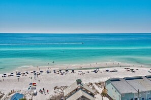 Ein Strand mit einem Steg, der ins Wasser ragt, feiner Sand, Sonnenschirme und Menschen, sowie Gebäude im Vordergrund.