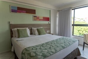 A hotel room with a large bed, a headboard with a wooden panel, two green pillows, a white bedspread, and a green patterned throw. There are two abstract paintings on the wall and a window with curtains. - Beautiful Room Near Cabo Branco Lighthouse - Beachfront Retreat with Ocean Views (Paraíba)
