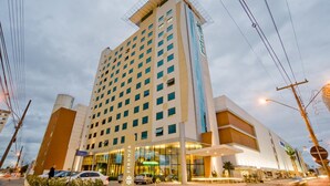 Um hotel com vários andares, uma entrada imponente, fachada de grandes vidros e uma estrutura no terraço.