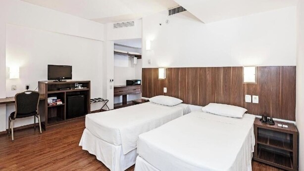 Quarto de hotel com duas camas, uma escrivaninha com televisão, uma cadeira e uma pequena escrivaninha com computador.