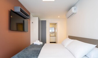 A hotel room with a bed, a flat-screen TV mounted on the wall, a built-in air conditioning unit, and a bathroom visible through an open door.
