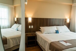 A hotel room with two beds, wooden headboards, bedside lamps, a small wooden nightstand, and a mirror. - Prime Location in Palhoça | Work-Friendly Unit | Pets Allowed (Santa Catarina)