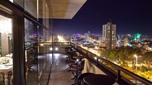 Ein Balkon mit Tischen und Stühlen, Blick auf die Stadt und ein Gebäude mit beleuchteten Fenstern.