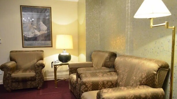 Ein Zimmer mit einer gemusterten Couch, zwei Sesseln, einem kleinen Beistelltisch mit einer Lampe und einem gerahmten Bild an der Wand.