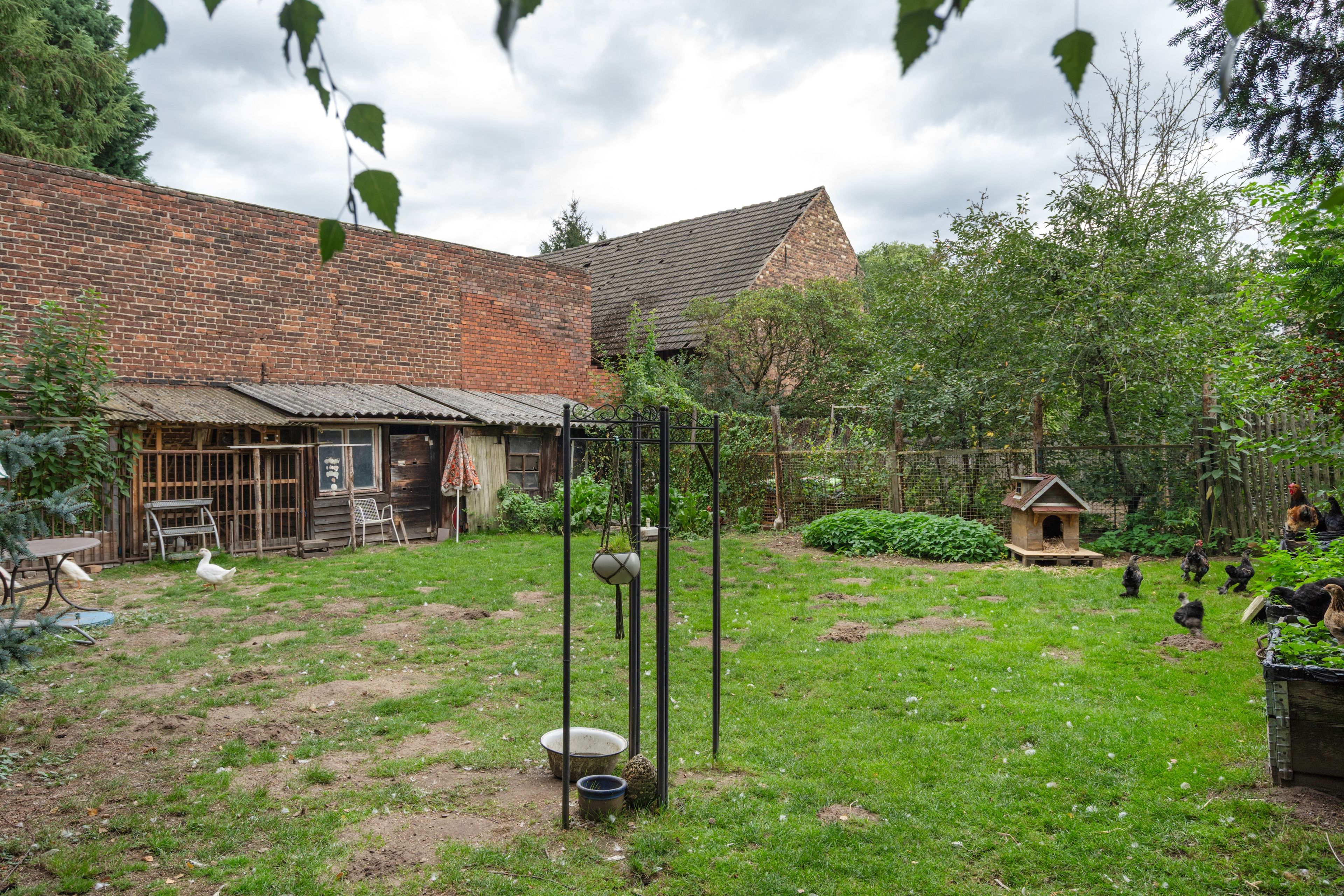 Un bâtiment rustique en briques, doté d’un toit en pente, entouré d’une cour verdoyante avec un poulailler et un petit jardin.