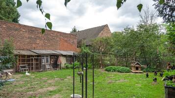 Un bâtiment rustique en briques, doté d’un toit en pente, entouré d’une cour verdoyante avec un poulailler et un petit jardin.