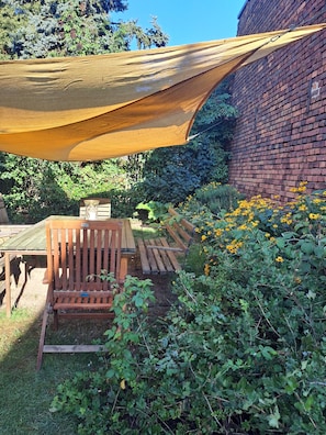 Een tuin met een houten bankje, een bakstenen muur en een geel zonnescherm.