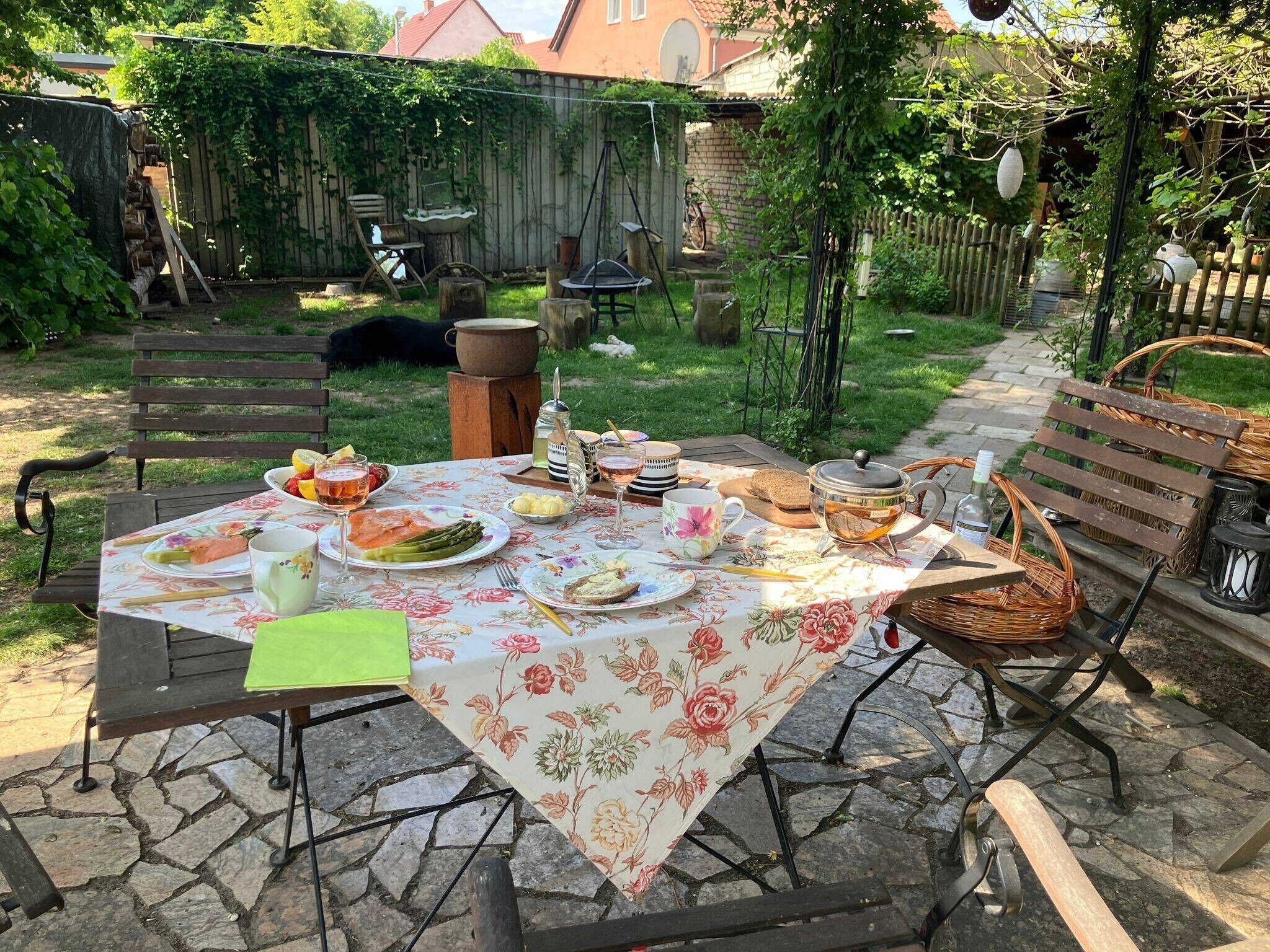 An outdoor dining area with a table set for a meal, surrounded by a garden and a wooden fence.
