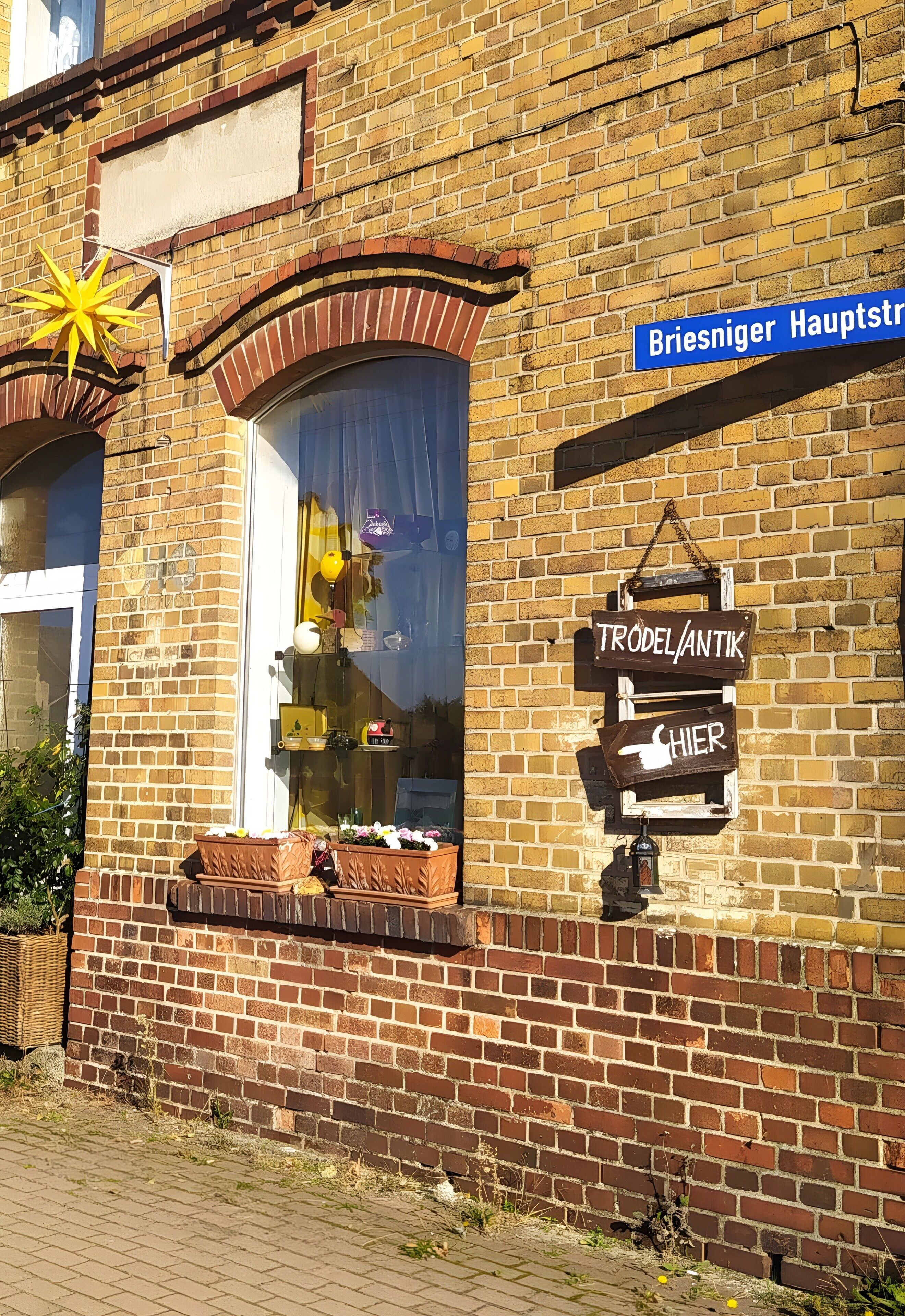Ein Backsteinhaus mit einem Schaufenster, das Waren ausstellt, einem Straßenschild mit der Aufschrift 'Briesniger Hauptstr' und einem Schild für 'TRODELANTIK'.
