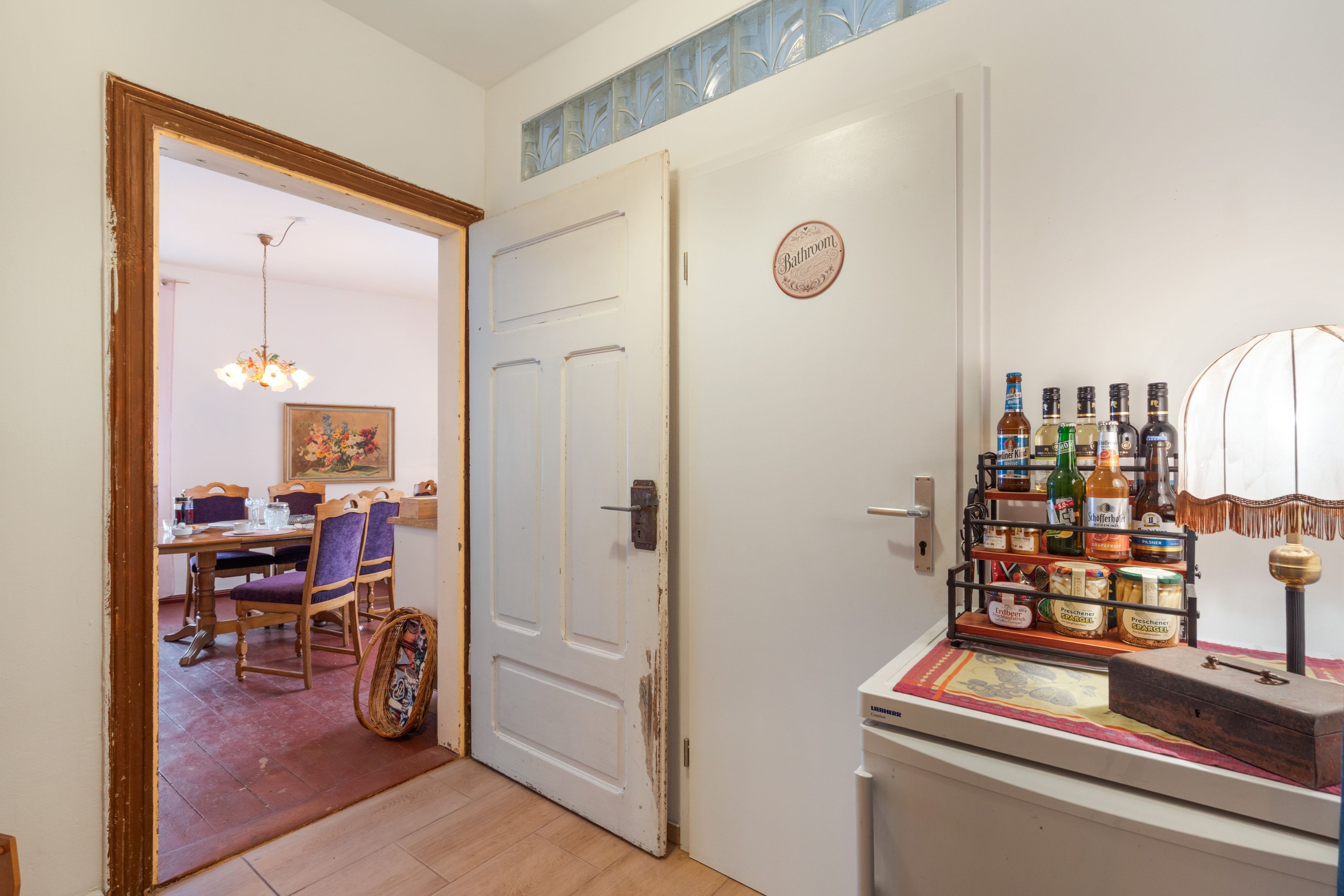 Una habitación con comedor, una puerta con el letrero 'Bathroom' y un estante con botellas y frascos varios.