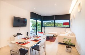 Een moderne woonkamer met een eethoek, een flatscreen-tv en grote ramen met uitzicht op groen.