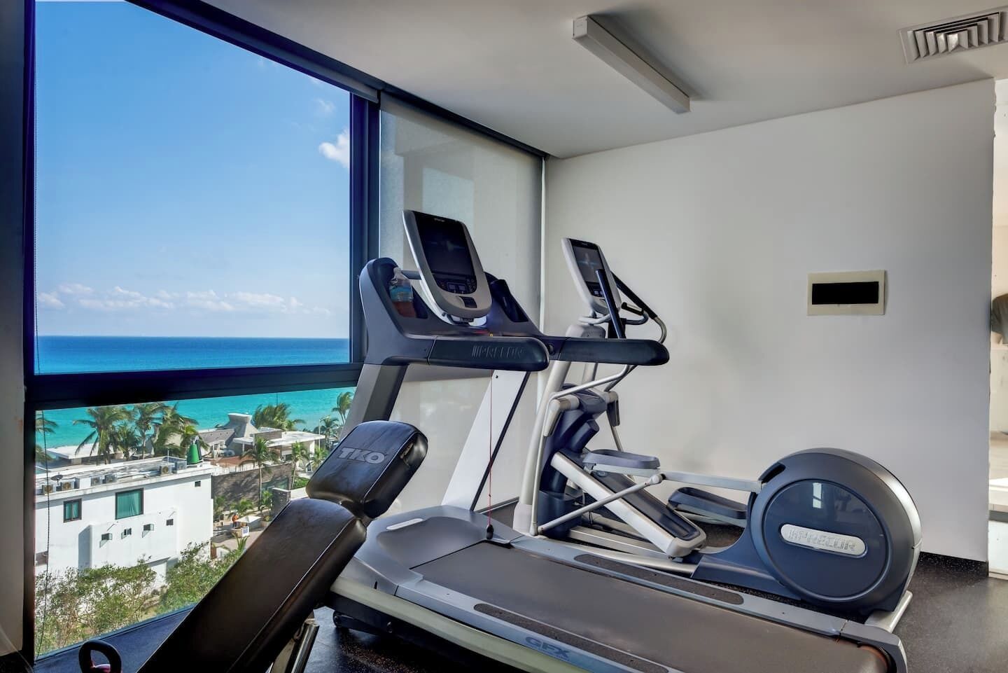 Un gimnasio con cintas de correr y una bicicleta estática, una ventana con vista al océano y una rejilla en el techo.