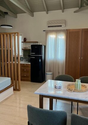 Een kleine kamer met een eettafel, stoelen, een koelkast en een houten garderobe.
