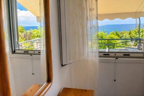 Quarto com janela que dá para uma varanda com vista para o mar e vegetação.