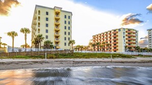 Ein Strandhotel mit mehreren Etagen, ein Sandstrand im Vordergrund und Palmen.