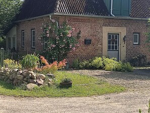 A brick house with a thatched roof, a front door, and a small garden with a stone wall and potted plants. - "Knight’s Room" in Farmhouse on Riding Estate (Malente)