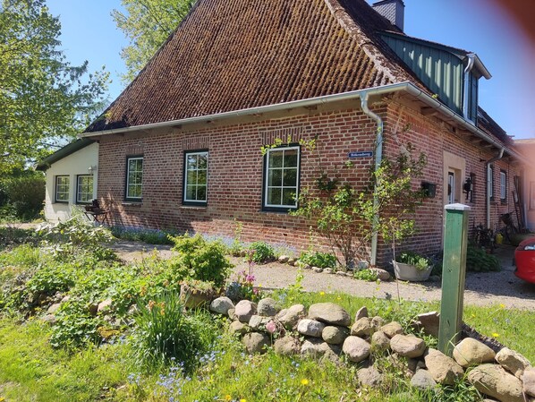 A brick building with a steeply pitched roof, a sign reading 'Bokensberg,' and a garden with rocks and flowering plants. - "Knight’s Room" in Farmhouse on Riding Estate (Malente)
