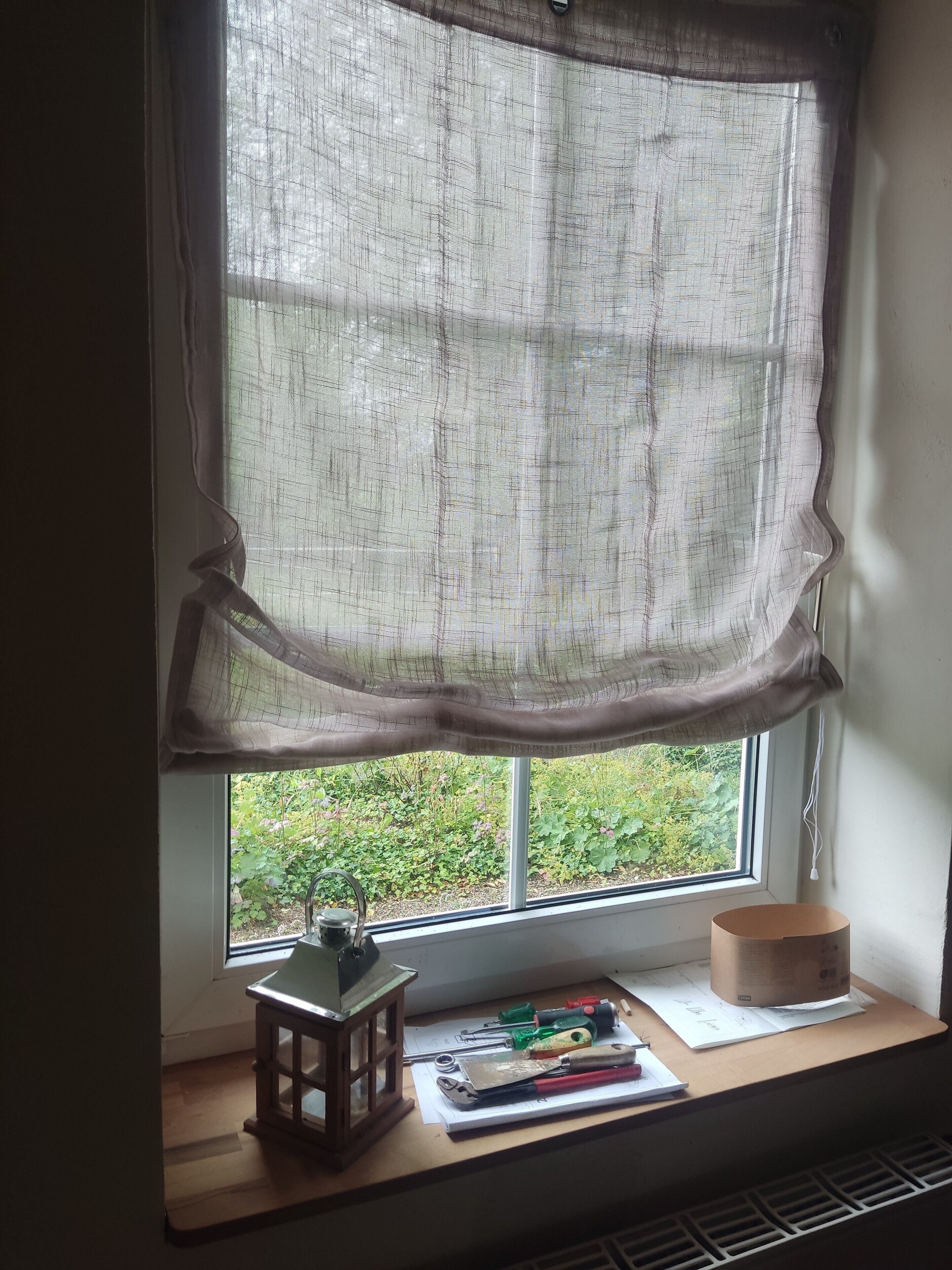 Ein Fenster mit einem durchsichtigen Vorhang, einer kleinen Holzlaterne und verschiedenen Werkzeugen auf einem Sims.