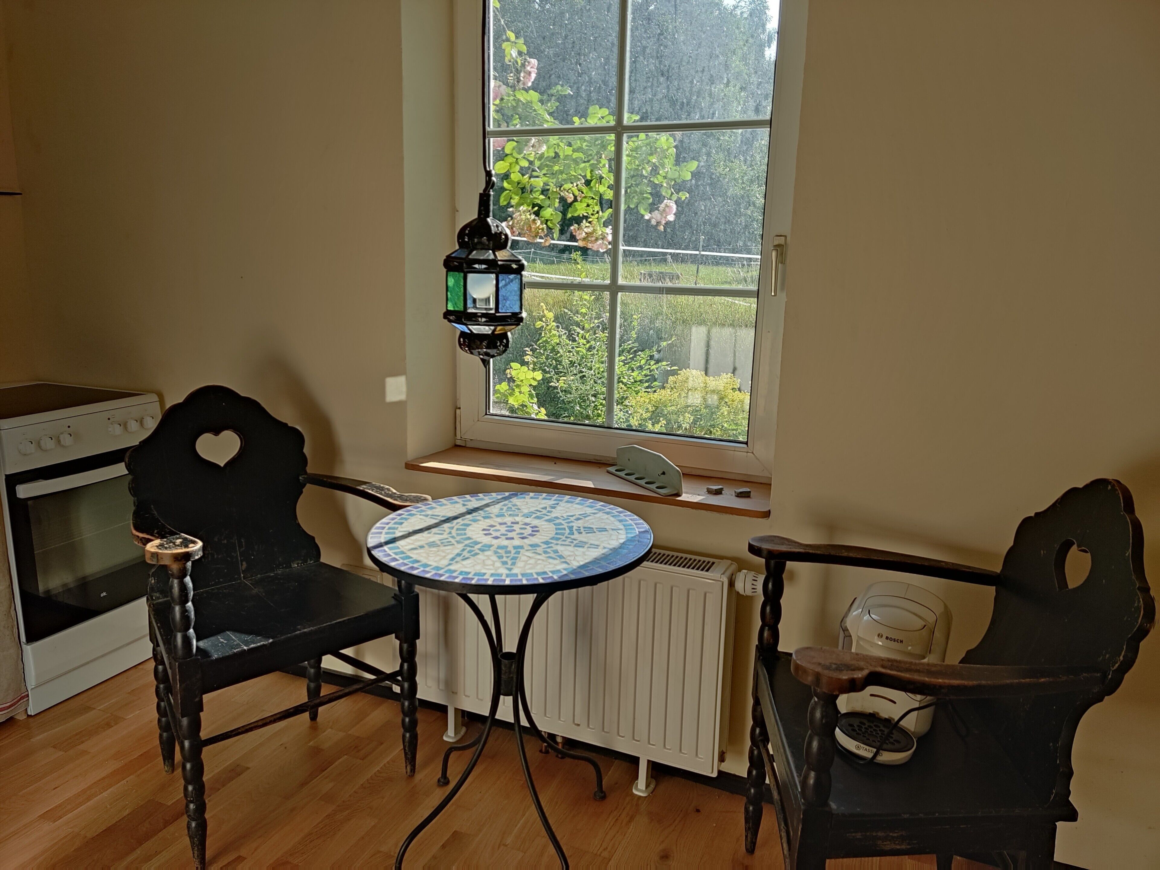 Eine Küche mit Herd, zwei Stühlen, einem Tisch und einem Fenster mit Blick auf Grünfläche.
